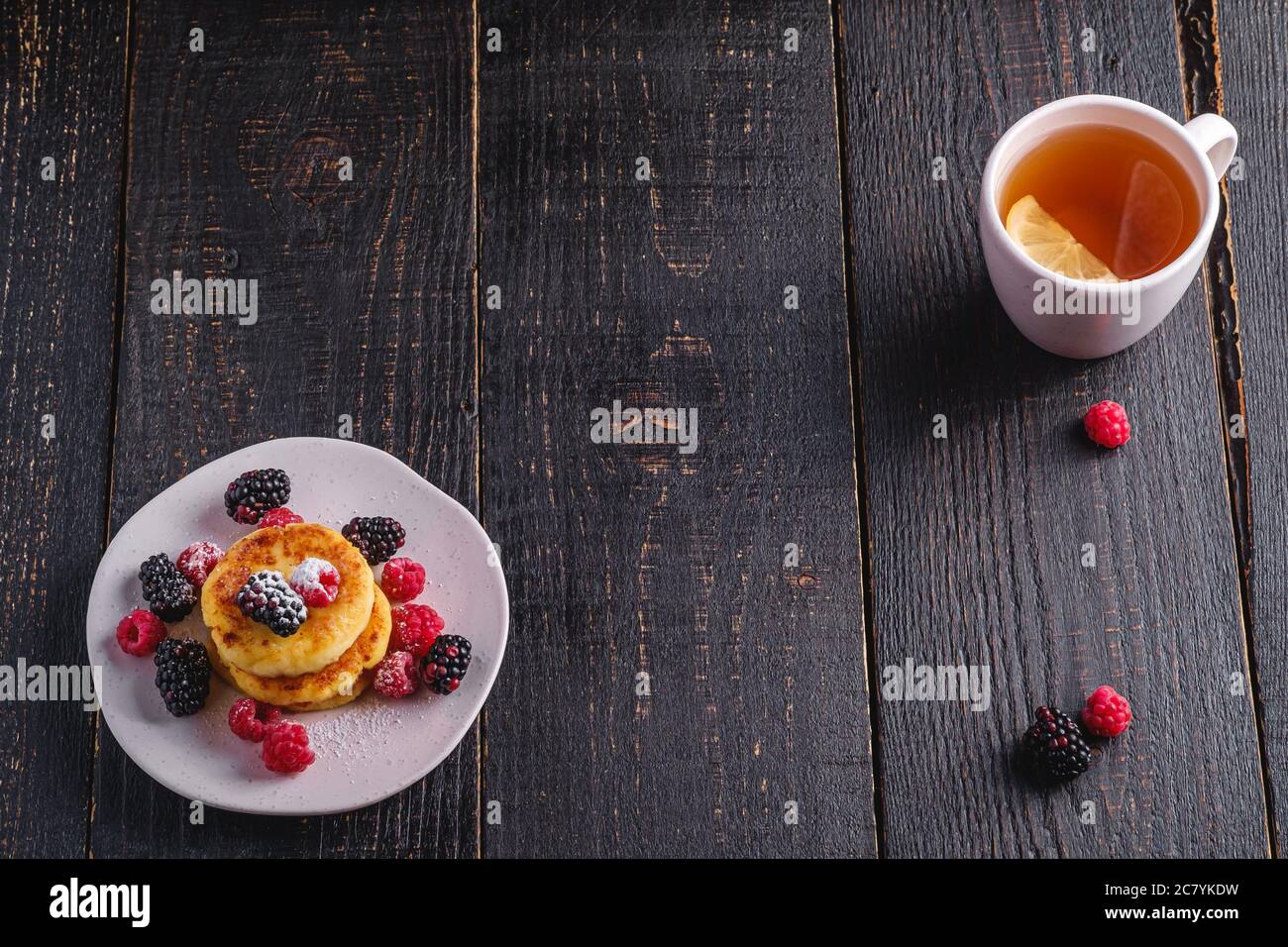 Frittelle di formaggio cottage, frittelle di cagliata dessert con lampone e frutti di bosco in piatto vicino alla tazza di tè caldo con fetta di limone Foto Stock