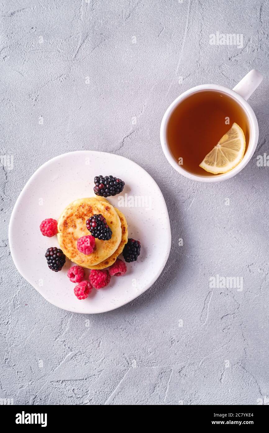 Frittelle di formaggio cottage, frittelle di cagliata dessert con lampone e frutti di bosco in piatto vicino alla tazza di tè caldo con fetta di limone Foto Stock