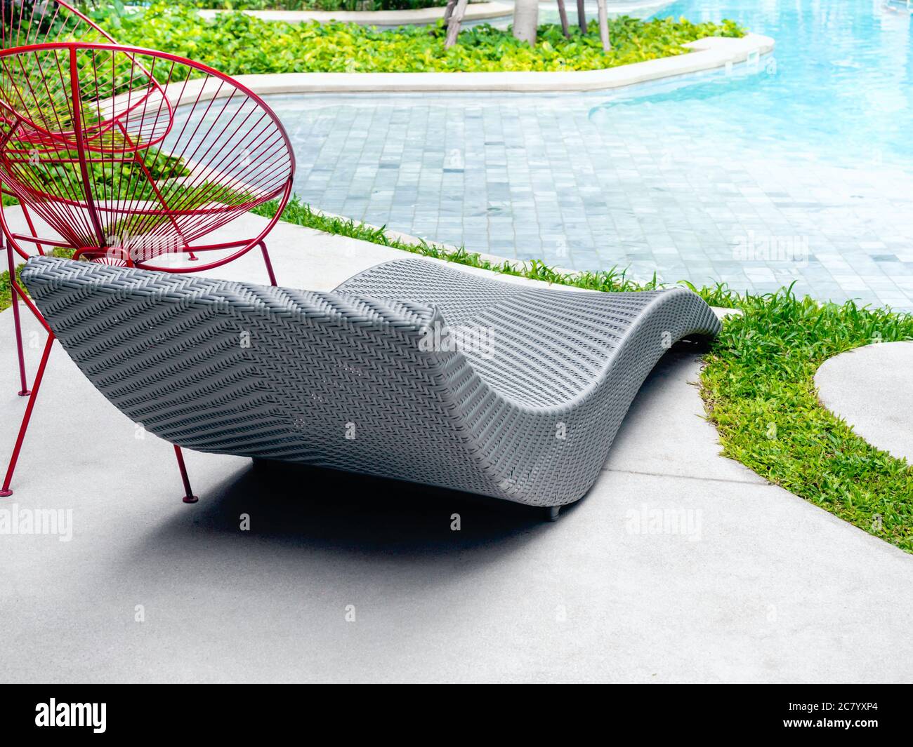 Moderno lettino ricurvo in vimini grigio e sedia in ferro rosso vicino alla piscina esterna e al giardino verde. Foto Stock