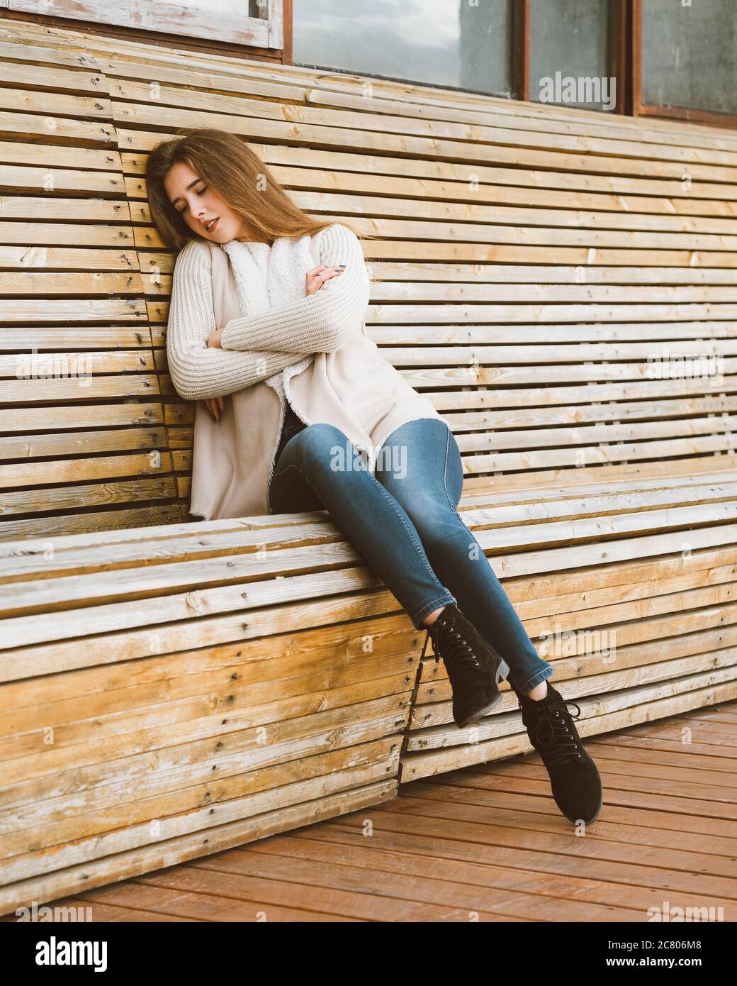 Bella ragazza giovane con lunghi capelli marroni si siede su panca di legno fatta di tavole e poggia, dorme o schizza in aria fresca. Scatta foto all'aperto con Foto Stock