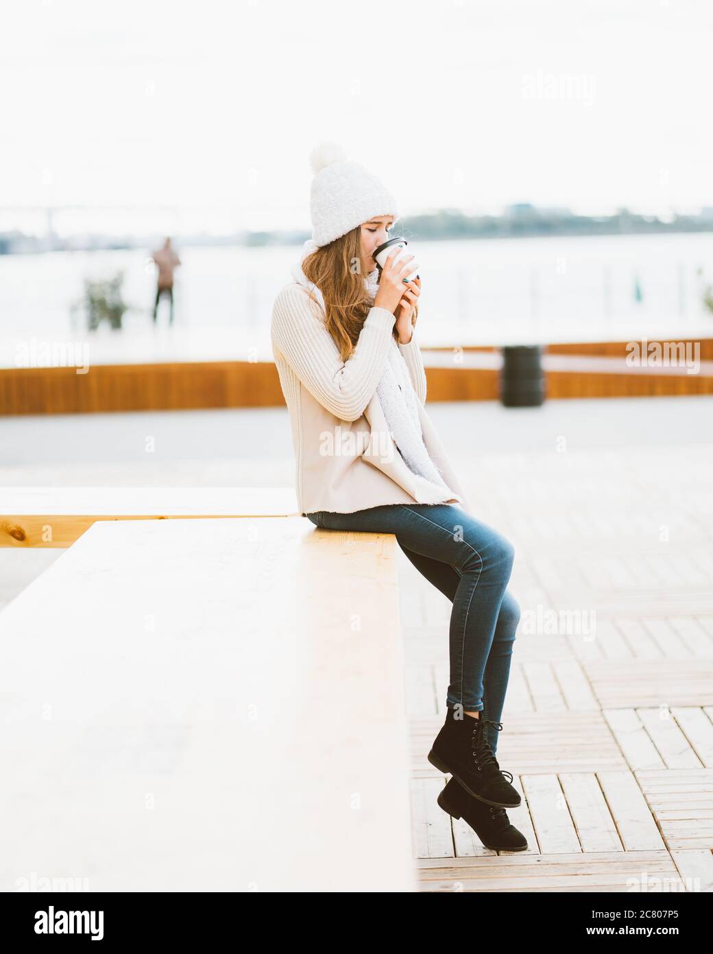 Bella giovane ragazza che beve caffè, tè da tazza di plastica in autunno, inverno. Una donna con capelli lunghi seduta sul lungomare sul Mar Baltico nel porto e. Foto Stock
