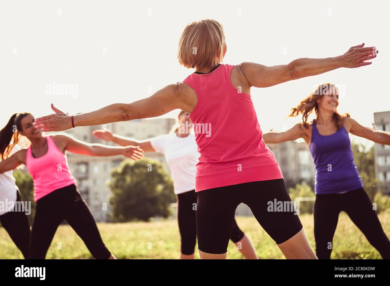Gruppo di giovani amici femminili che si allenano in un parco o campo all'aperto ridendo e sorridendo in una vista da dietro la donna che guida la classe in un healt Foto Stock