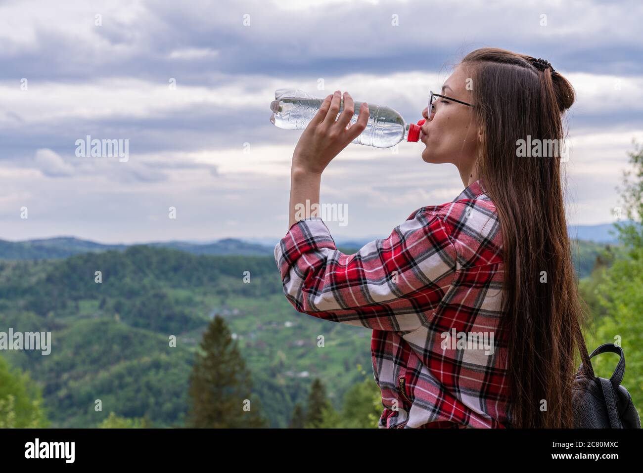 Una ragazza di aspetto europeo con capelli sciolti brunette contro un cielo nuvoloso e montagne. Beve acqua da una bottiglia. Stile di vita sano, atletico Foto Stock