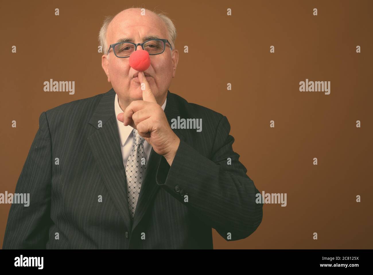 Uomo d'affari anziano in sovrappeso che indossa il naso di clown su sfondo marrone Foto Stock