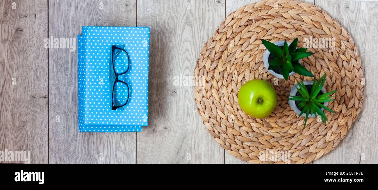 Torna a scuola, mucchio di libri in coperte blu, bicchieri, mela verde e piante in vasi su un vimini stand su tavolo di legno. Istruzione domestica di distanza. Quar Foto Stock