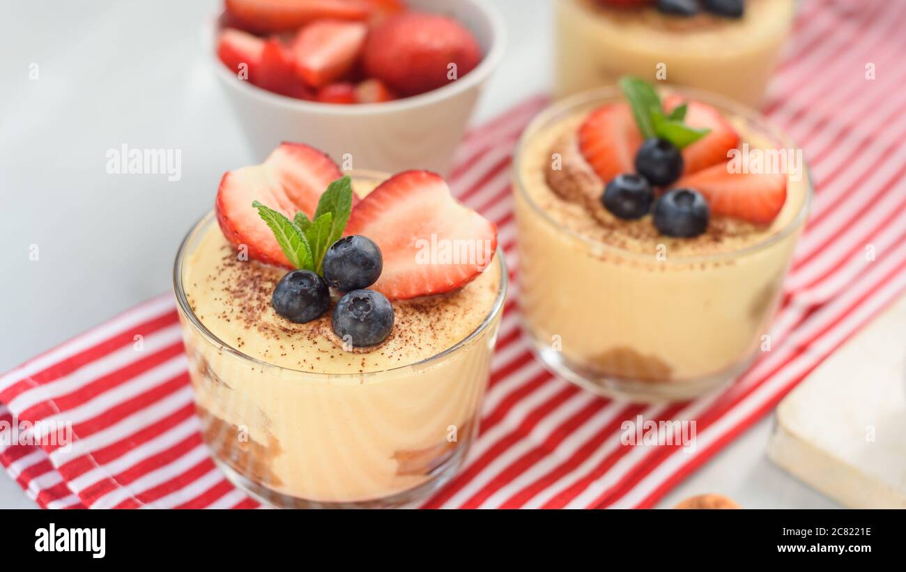 tiramisù, squisito dessert fatto in casa in bicchieri decorati con fragola, mirtillo, menta su tavolo di legno bianco, vista dall'alto. Foto Stock
