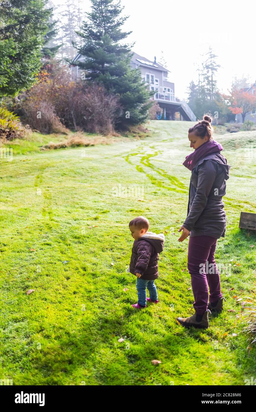 Madre con figlia giovane in erba in un cortile dietro una casa; Whidbey Island, Washington, Stati Uniti d'America Foto Stock