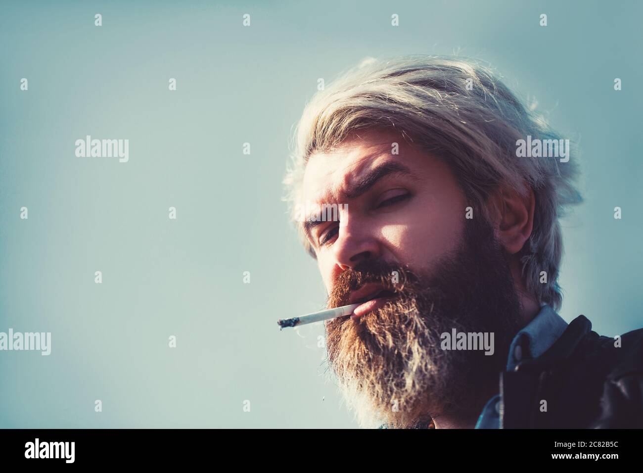 Uomo serio bearded che fuma una sigaretta sulla spiaggia. Hipster bearded sul cielo. Ritratto in primo piano. Foto Stock