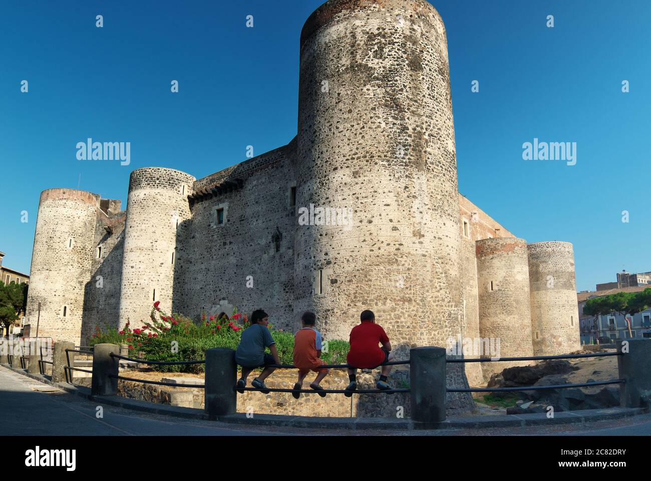 Tre bambini sono seduti sulla ringhiera di fronte al Castello Ursino di Catania, simbolo dell'architettura medievale siciliana Foto Stock
