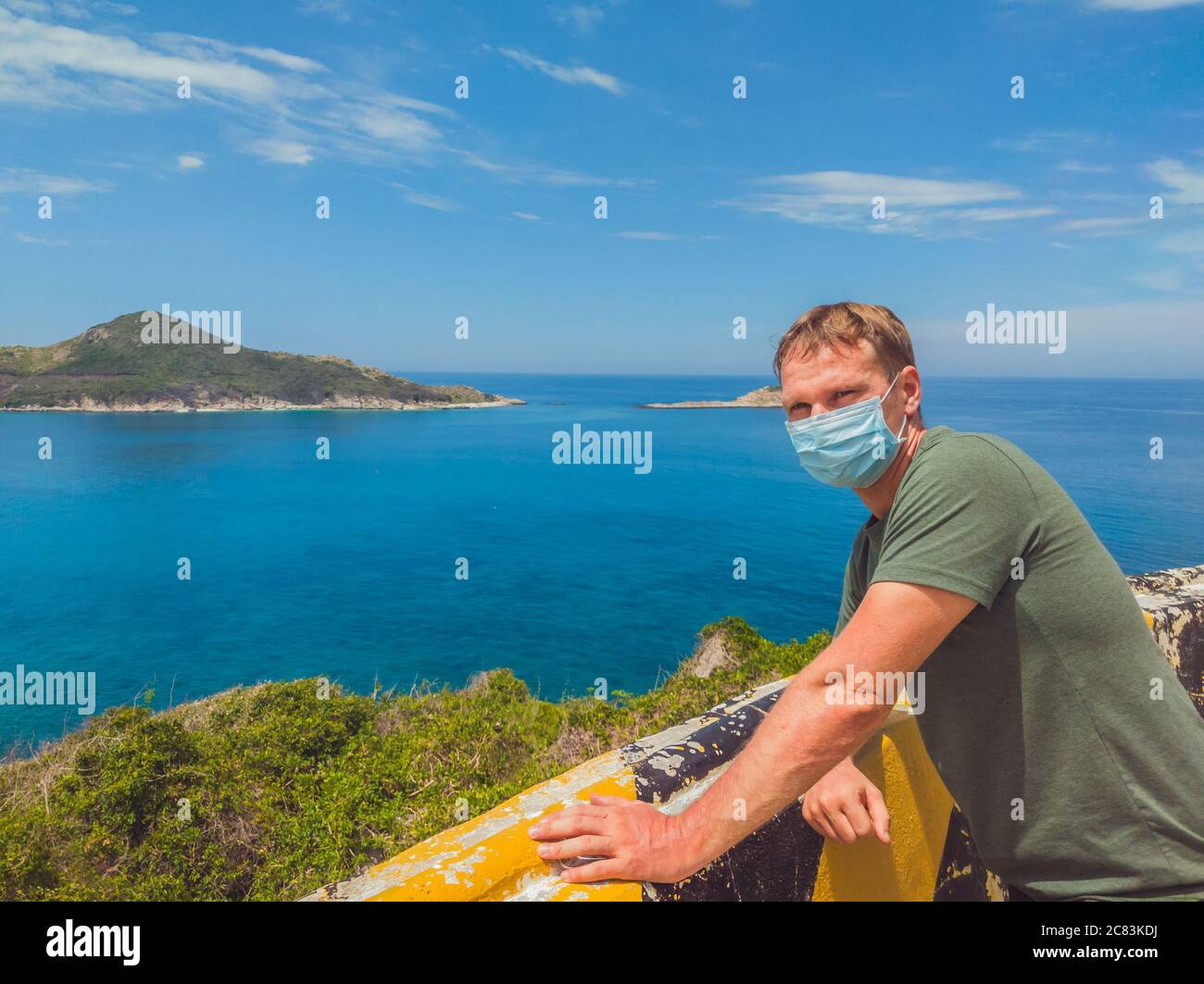 Giovane uomo bello proteggere da contaminazione aria o coronavirus indossando maschera medica, a piedi all'esterno nel parco vicino mare azzurro, cielo blu. Stop Foto Stock