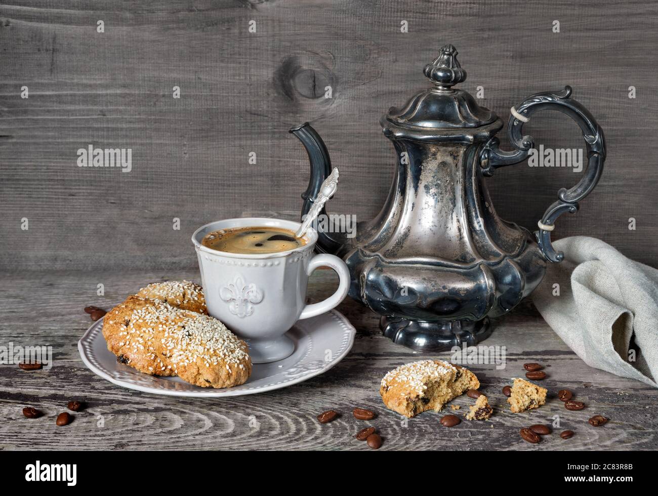 Caffè nero in una tazza vittoriana stilizzata e biscotti d'avena cosparsi di semi di sesamo su un piatto di porcellana, oltre a una vecchia caffettiera argentata e gra Foto Stock
