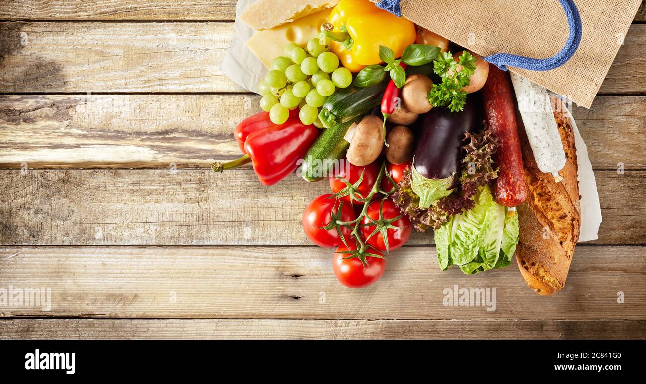 Ingredienti freschi di cucina che si riversano da una borsa con verdure assortite, uva, salsicce piccanti e baguette croccante su un panorama rustico in legno Foto Stock