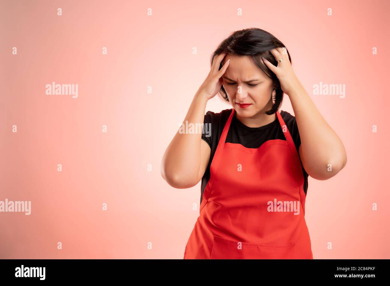 Donna impiegata al supermercato con grembiule rosso e maglietta nera, ha mal di testa isolato su sfondo rosso Foto Stock