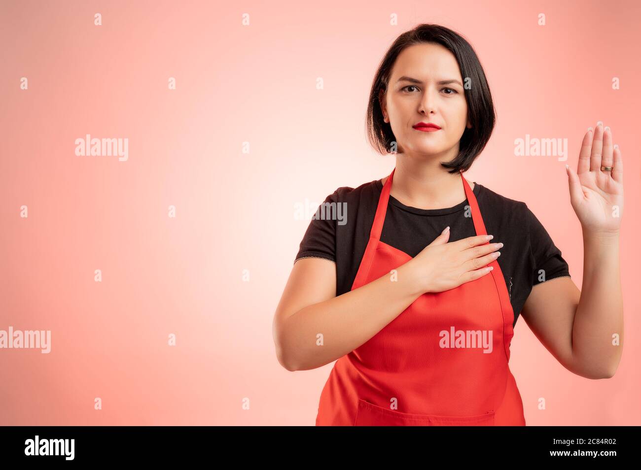 Donna impiegata al supermercato con grembiule rosso e maglietta nera, che mostra giuramento isolato su sfondo rosso Foto Stock