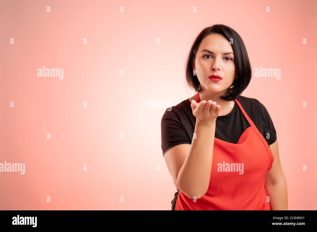 Donna impiegata al supermercato con grembiule rosso e t-shirt nera, inviare baci isolati su sfondo rosso Foto Stock