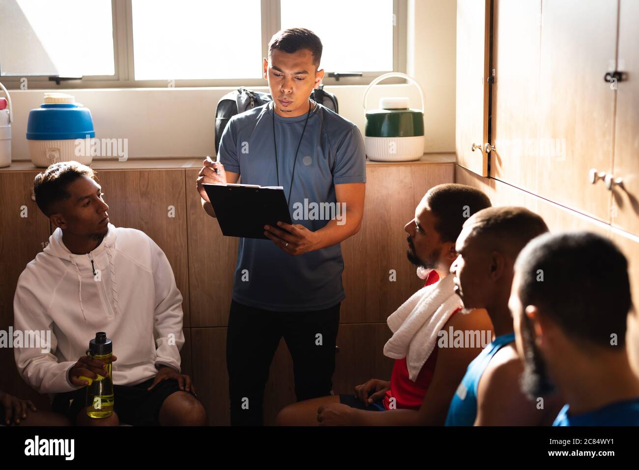 Gruppo multietnico di giocatori di calcio che indossano abiti sportivi seduti in spogliatoio durante una pausa di gioco, ascoltando il loro allenatore che istruisce ho Foto Stock