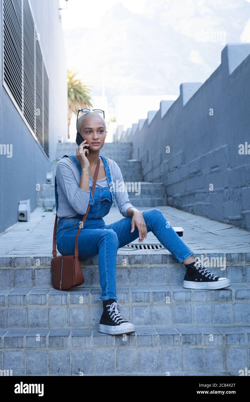 Misto razza alternativa donna con capelli biondi corti fuori e circa in città in una giornata di sole, indossando occhiali da sole e denim dungarees, seduto su gradini Foto Stock