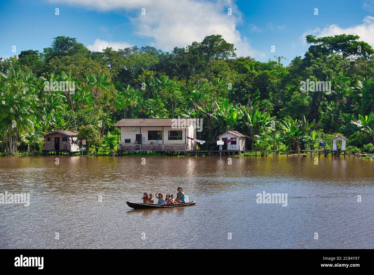 Un adulto e bambini in una canoa di legno aperta a. uno sfondo di case e capanne sulle rive del Il fiume Amazzonia con la foresta pluviale oltre in Brasile Foto Stock