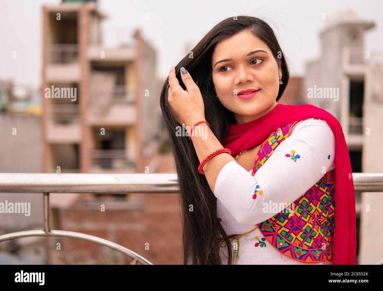 Bella giovane donna indiana che tiene i suoi lunghi capelli neri e in piedi vicino alla ringhiera sul tetto. Sta guardando via e pensando con sorriso. Foto Stock