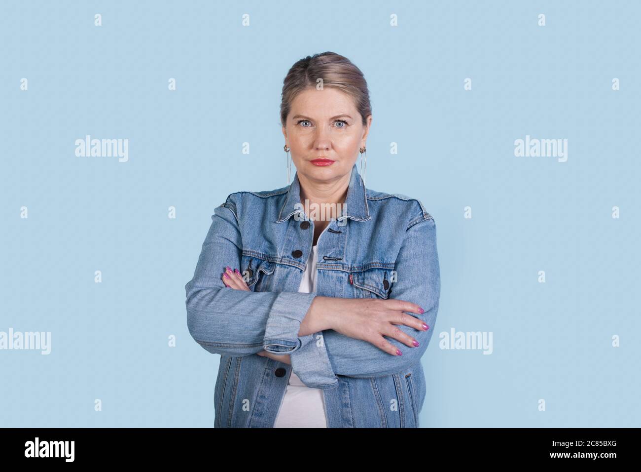 Donna caucasica anziana seria con capelli biondi che indossa cappotto di jeans che posa con le mani incrociate su una parete blu dello studio Foto Stock