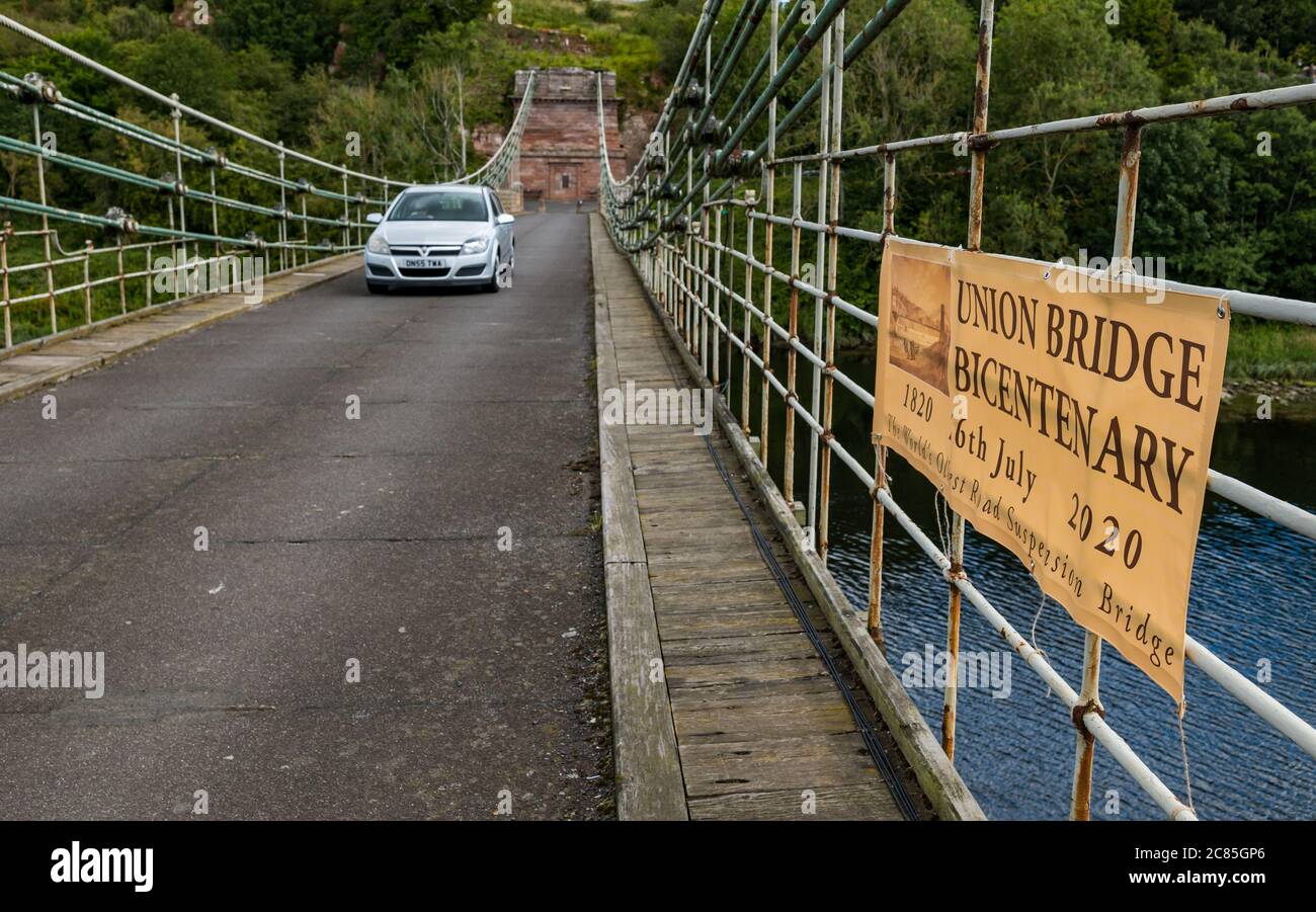 River Tweed, confine inglese/scozzese, Regno Unito, 21 luglio 2020. Bicentenario di Union Bridge: Il ponte celebra il suo 200° anniversario il 26 luglio. Fu il primo ponte sospeso veicolare nel Regno Unito. Al momento della sua costruzione, era il ponte sospeso in ferro battuto più lungo del mondo a 137 m. È ancora utilizzato regolarmente da pedoni, ciclisti e auto, ma le auto devono attraversare in un momento. Questa vista è dal confine scozzese. Un banner annuncia la celebrazione del bicentenario mentre un'auto attraversa il ponte Foto Stock