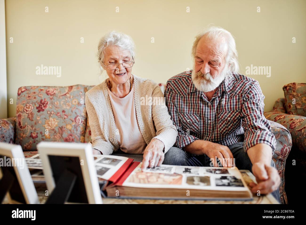 Uomo anziano seduto sul divano con la sua moglie anziana che guarda le foto all'albuum di famiglia, ricordando momenti significativi della vita Foto Stock