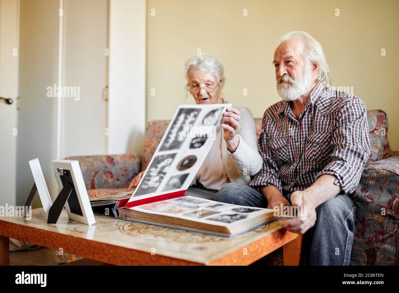 Coppia senior guardando un album fotografico di famiglia con foto d'epoca seduto sul divano in soggiorno con interni morbidi e accoglienti, la vecchia signora punta alle foto Foto Stock