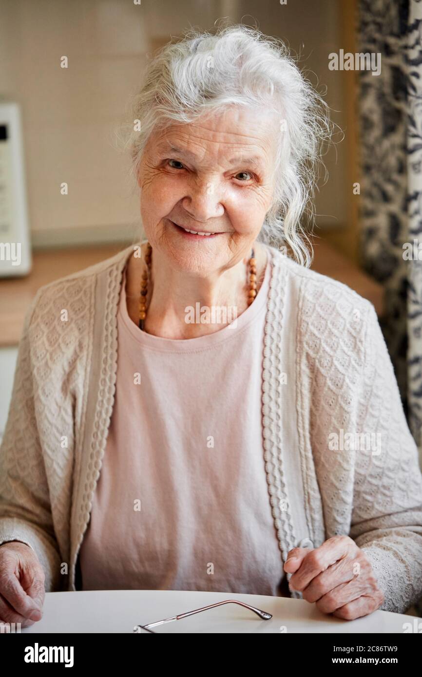 Primo piano ritratto di donna anziana con viso strinlke e capelli grigi, sorridente e seduto al tavolo a casa, concetto di gente di età avanzata Foto Stock
