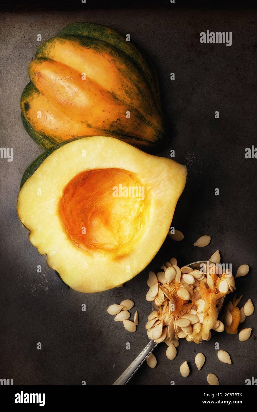 Immagine piatta di una zucca di Acorn tagliata a metà su una lamiera da forno con un cucchiaio e semi scopati con luce laterale calda. Foto Stock