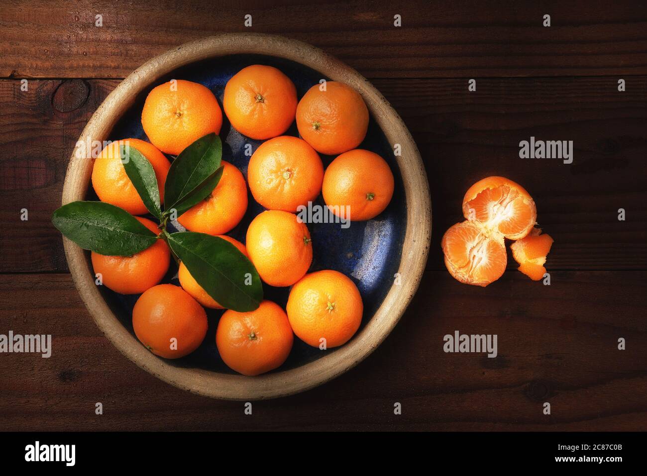 Immagine piatta di un piatto di arance mandarine fresche selezionate in un tavolo rustico in legno con un pezzo di frutta sbucciata con luce laterale calda. Foto Stock