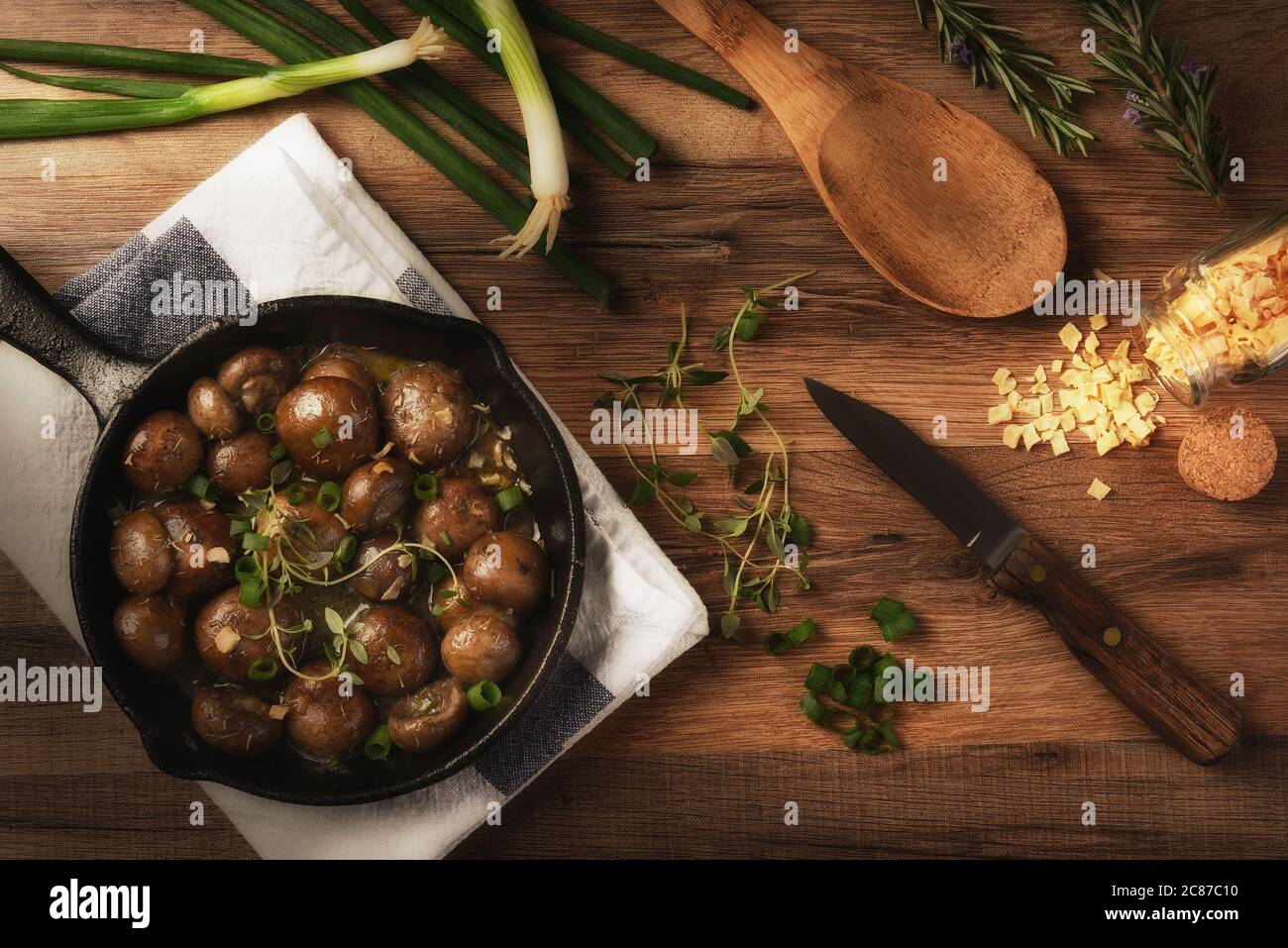 Vista piatta dei funghi saltati in una padella di ghisa circondata da ingredienti e utensili. Orizzontale su un tavolo rustico da cucina con lato caldo Foto Stock
