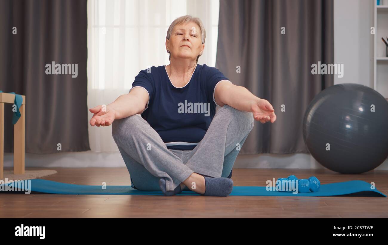 Donna anziana meditating seduta sul tappeto yoga in soggiorno. Attivo sano stile di vita sportivo vecchia persona allenamento home benessere e esercizio interno Foto Stock