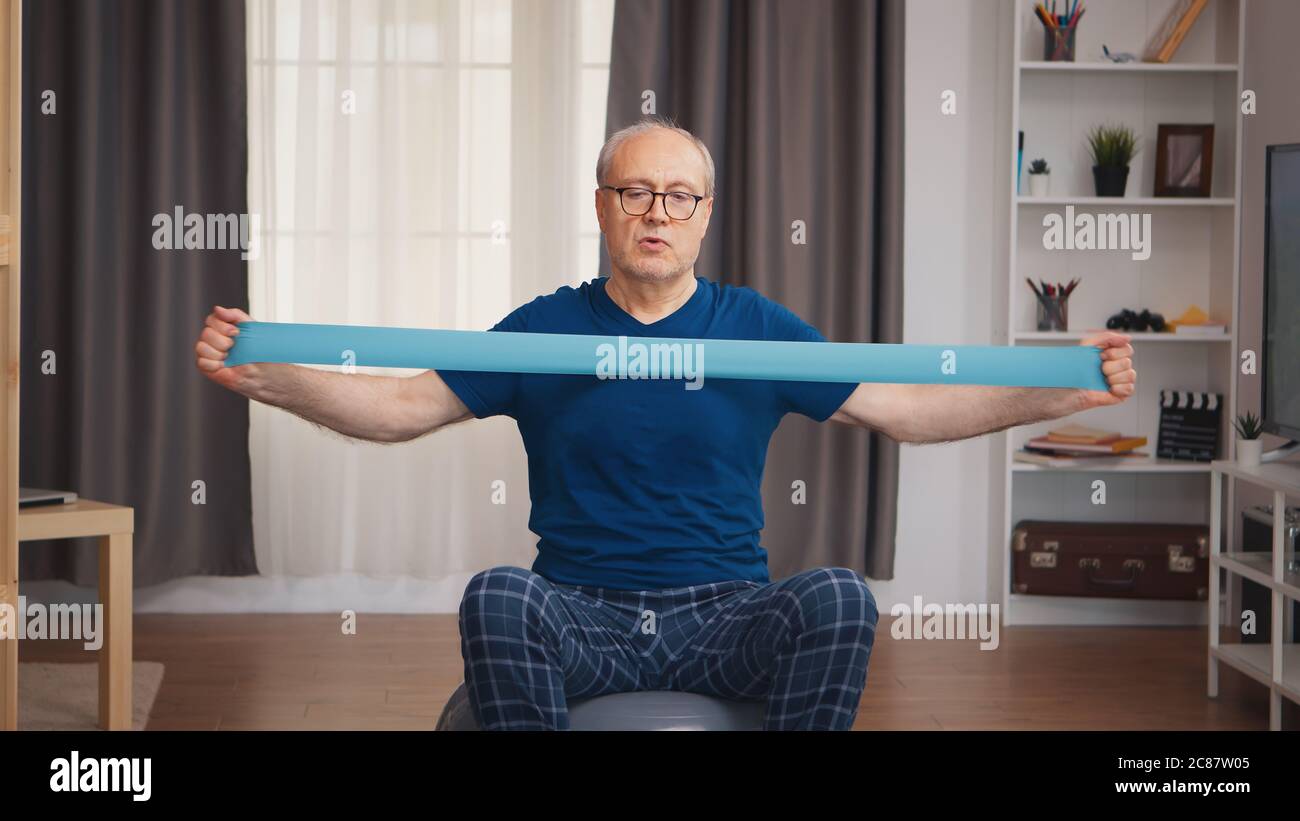 Uomo anziano sull'addestramento della sfera di equilibrio con la fascia di resistenza. Anziano pensionato sano addestrando sport di salute a casa, esercitandosi attività di idoneità all'età anziana Foto Stock