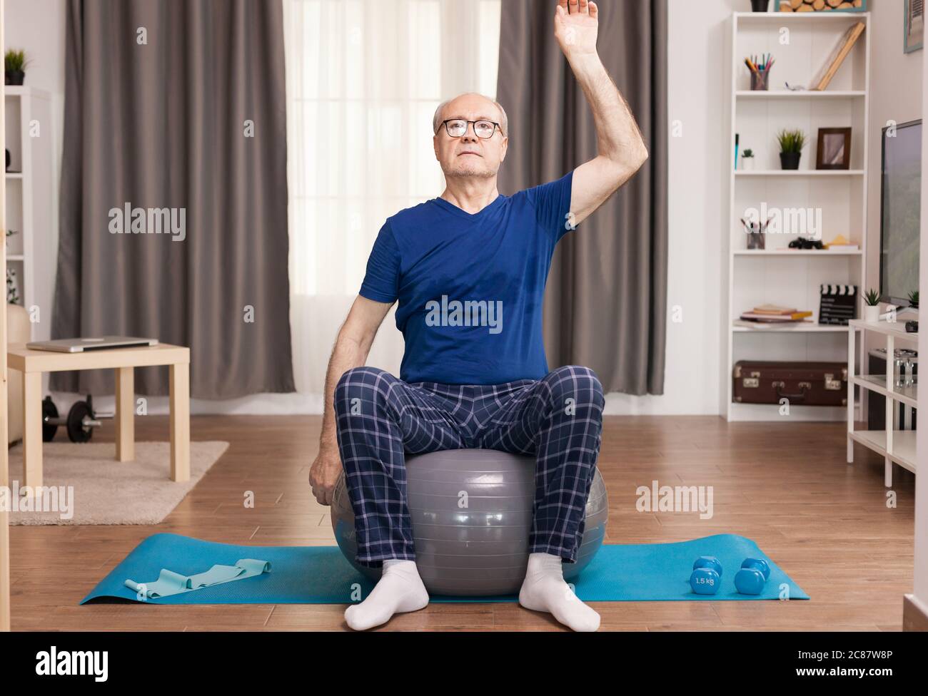 L'uomo anziano si alleducazione a casa, facendo esercizi al braccio. Anziano pensionato online internet esercizio di formazione a casa attività sportiva con manubri, banda di resistenza, palla svizzera all'età di anziano pensionamento. Foto Stock