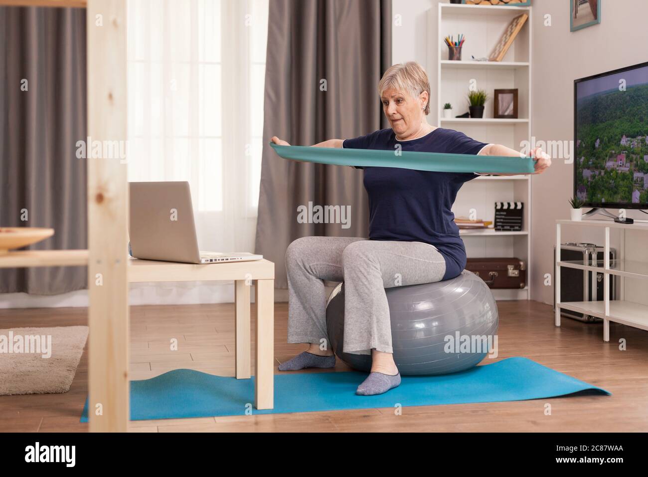 donna di 60 anni che si esercita da casa. Anziano pensionato online internet esercizio di formazione a casa attività sportiva con manubri, banda di resistenza, palla svizzera all'età di anziano pensionamento Foto Stock