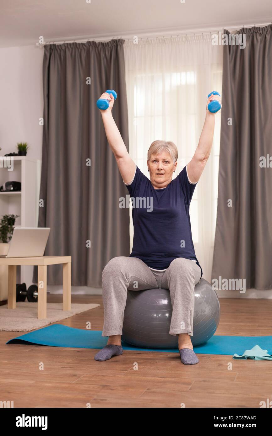 Donna anziana che si esercita con manubri per perdere peso. Anziano pensionato online internet esercizio di formazione a casa attività sportiva con manubri, banda di resistenza, palla svizzera all'età di anziano pensionamento Foto Stock