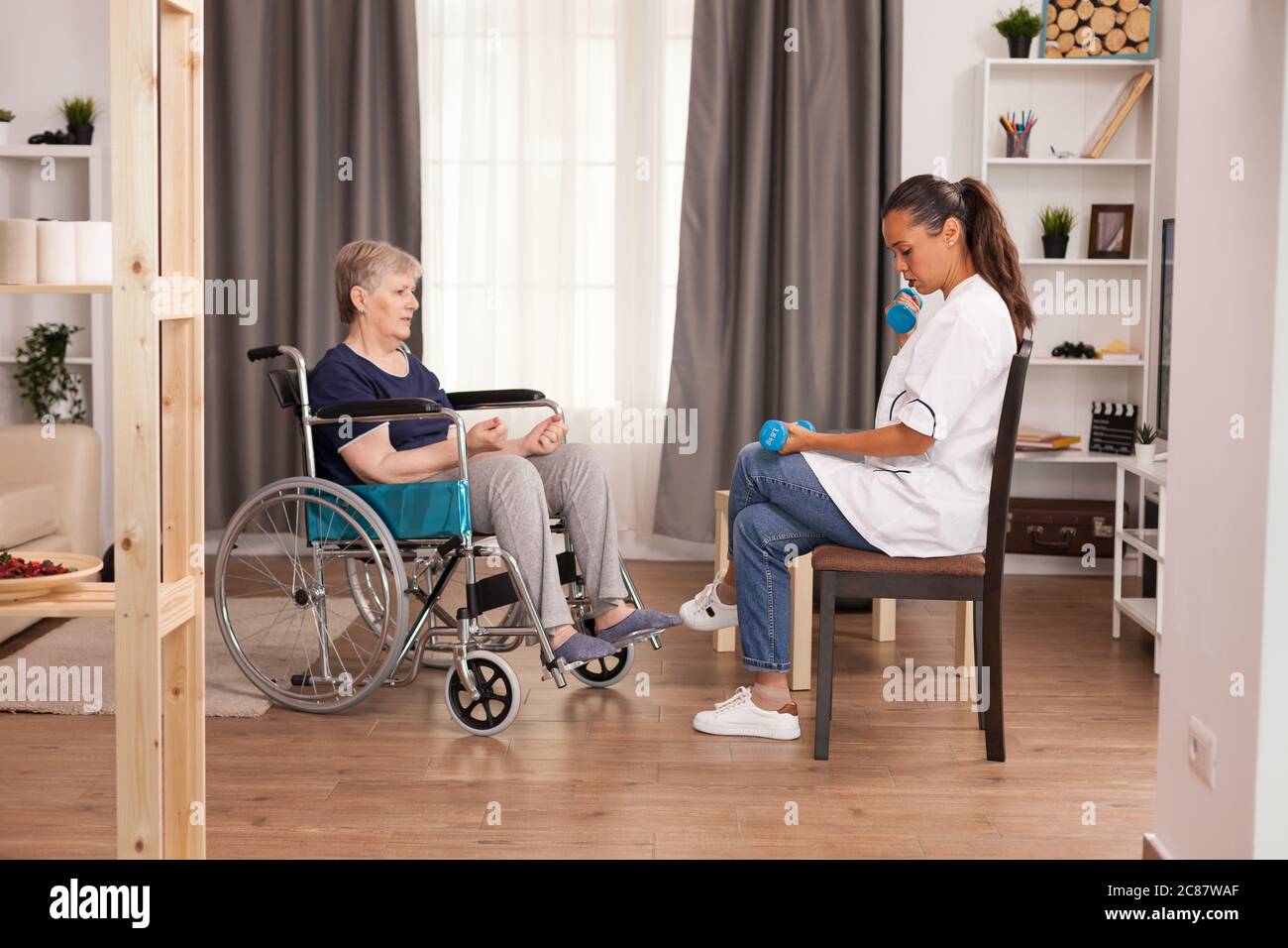 Medico che mostra ad esercizi immobilizzati di vecchia donna con manubri per il recupero fisico. Disabili anziani ricoverati infermiere professionista di aiuto, cura casa di riposo e riabilitazione Foto Stock