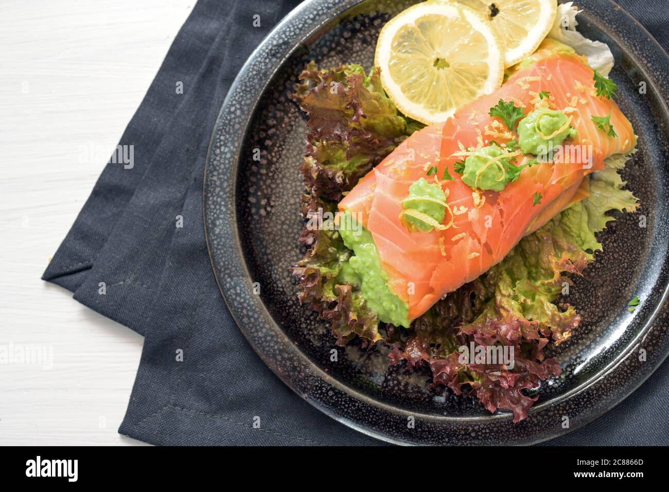 Salmone affumicato riempito con purea di piselli su insalata di lattuga con fette di limone e prezzemolo guarnire su un piatto scuro e tovagliolo, tavolo bianco, insalata di mucca sana o. Foto Stock