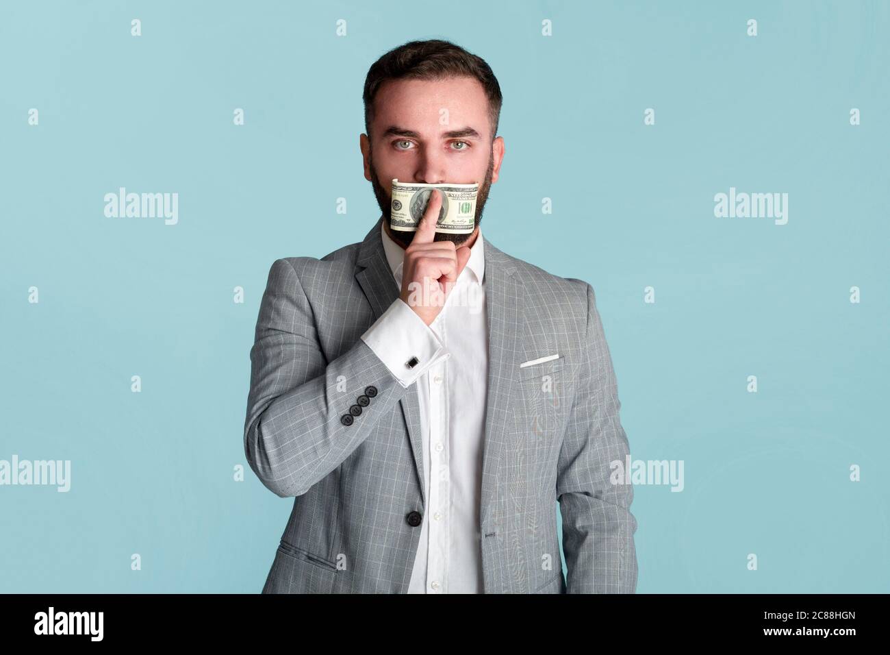 Silenziato dal denaro. Giovane uomo d'affari che copre la sua bocca con cento dollari di conto su sfondo blu Foto Stock