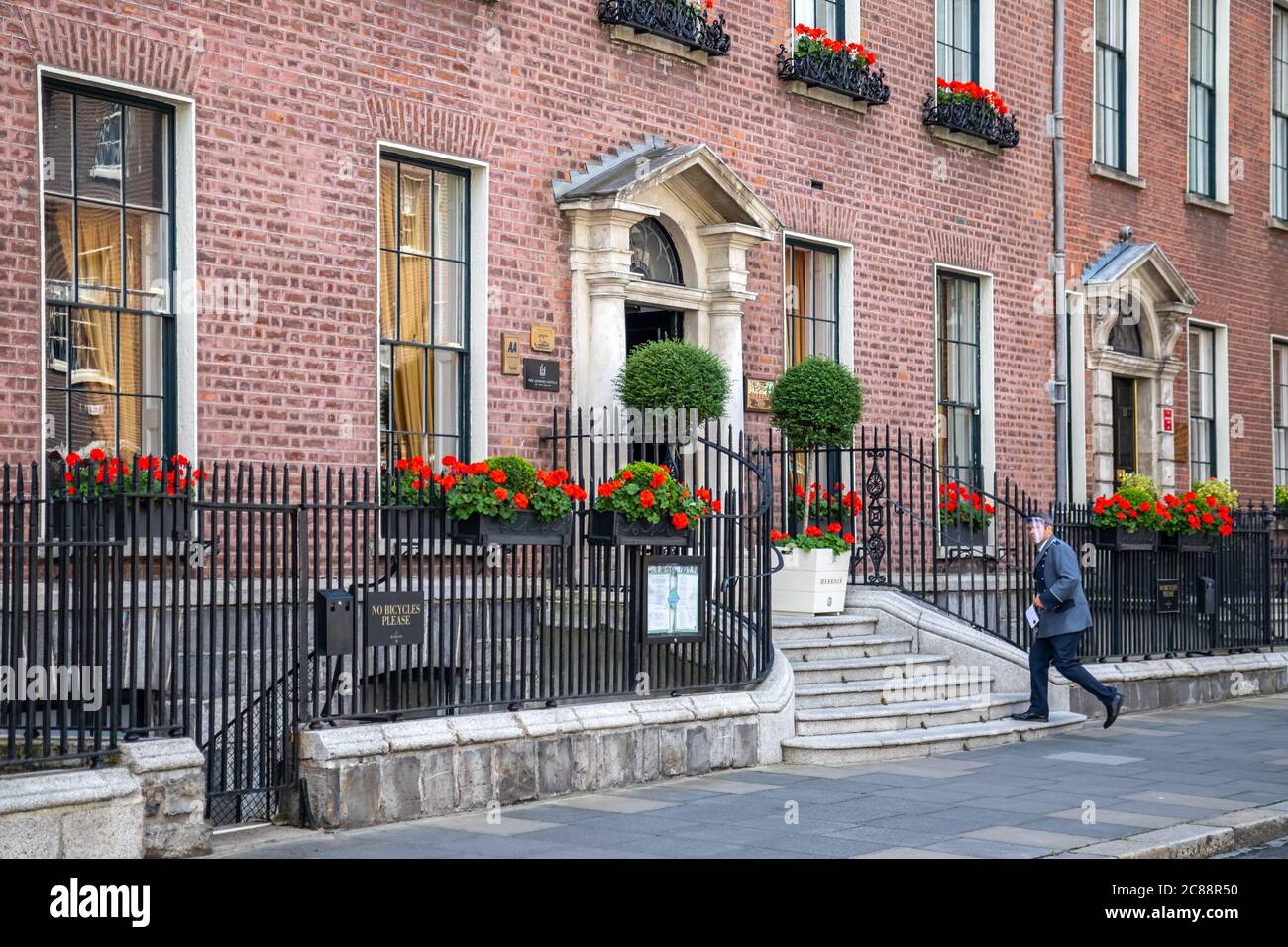 Ingresso frontale al 'The Merrion Hotel' con personale che indossa visiera protettiva durante la pandemia di Coronavirus Covid-19. Hotel gerani rossi Foto Stock