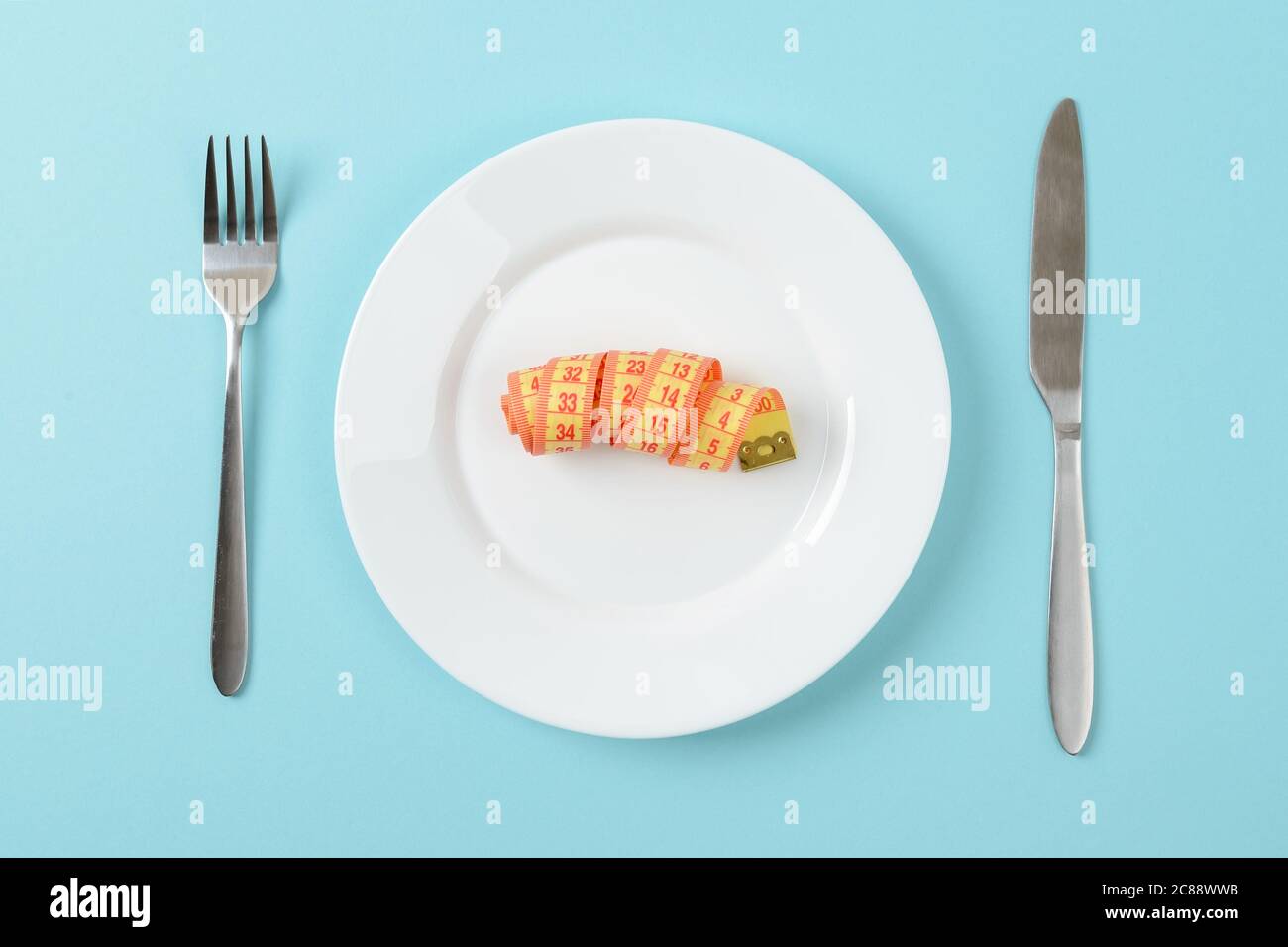 Rotolo di nastro di misurazione su un piatto da pranzo bianco, coltello da tavolo e forchetta su sfondo blu. Concetto di dimagrimento, controllo del peso corporeo e alimentazione sana Foto Stock