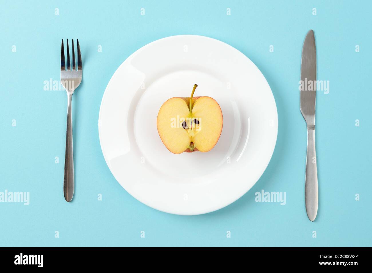 Metà di mela matura succosa su un piatto da pranzo bianco, coltello da tavola e forchetta su sfondo blu. Concetto di alimentazione sana, vegetarianismo, dieta di cibo crudo. Foto Stock