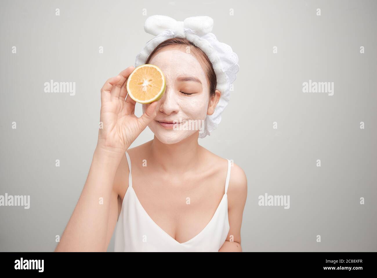 Giovane donna con maschera facciale in argilla che tiene una fetta di frutta arancione che copre gli occhi Foto Stock Giovane donna con maschera facciale in argilla che tiene una fetta di frutta arancione che copre gli occhi Foto Stock