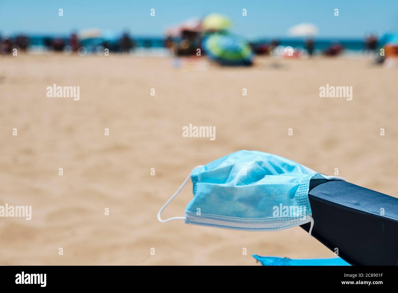 primo piano di una maschera chirurgica sul bracciolo di una sedia blu ponte sulla spiaggia, con l'oceano sullo sfondo Foto Stock