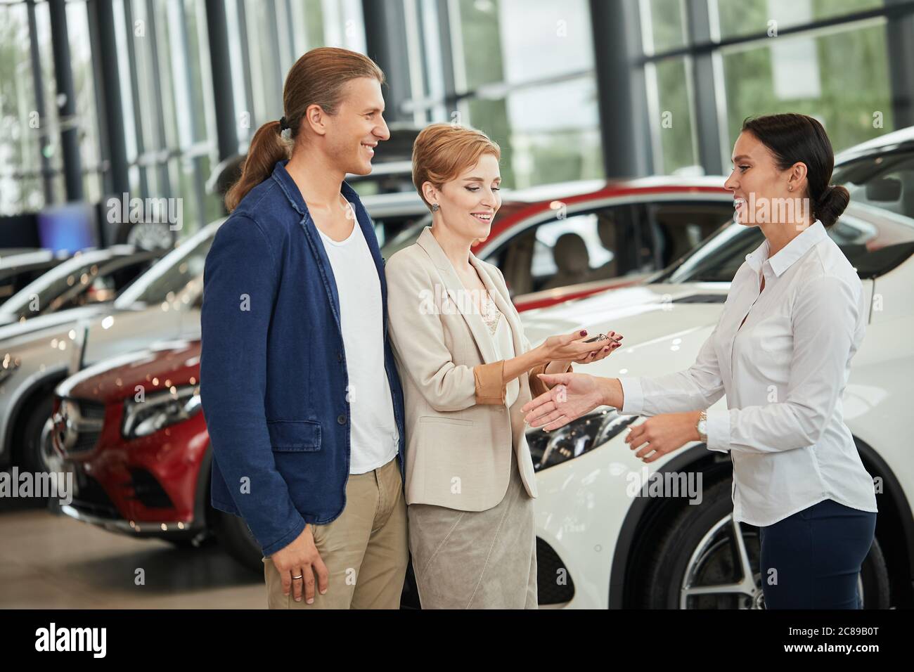 Proprietari di auto nuove. Felice giovane coppia bionda vestita di abbigliamento formale, ricevendo le chiavi di un venditore di auto presso la concessionaria. Foto Stock