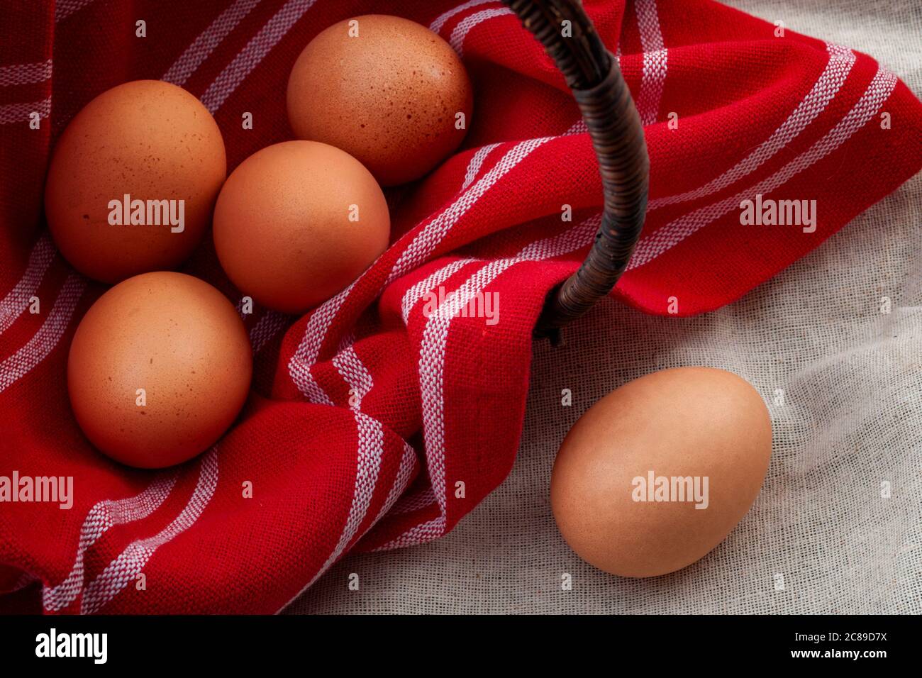 Ingredienti naturali, cibo sano e ambiente rustico con un cesto di vimini coperto da un panno rosso vintage capovolto su un lato con uova interne Foto Stock