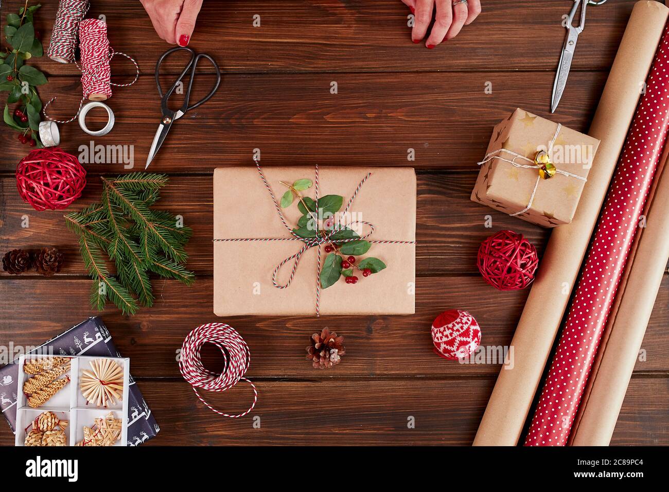 Piatto di Natale posa di scatola regalo artigianale non pronta su tavolo rustico in legno con rotoli di carta da imballaggio artigianale e a colori, coni di pino, rami di abete e. Foto Stock