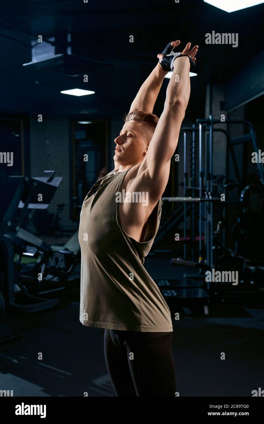 Bell'allestitore in abbigliamento sportivo che allunga la parte superiore del corpo nel club sportivo. Vista laterale di un sportivo caucasico in guanti che si posano in palestra vuota. Concetto di bodybuilding, stile di vita sano. Foto Stock