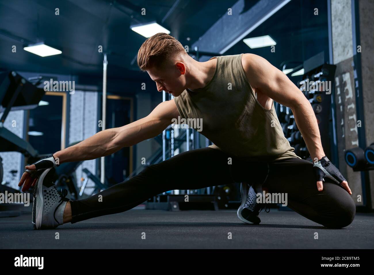 Allestitore caucasico muscolare in abbigliamento sportivo che si estende sul pavimento in club sportivo. Vista frontale della bella tomaia sportiva in palestra vuota. Concetto di bodybuilding, stile di vita sano. Foto Stock