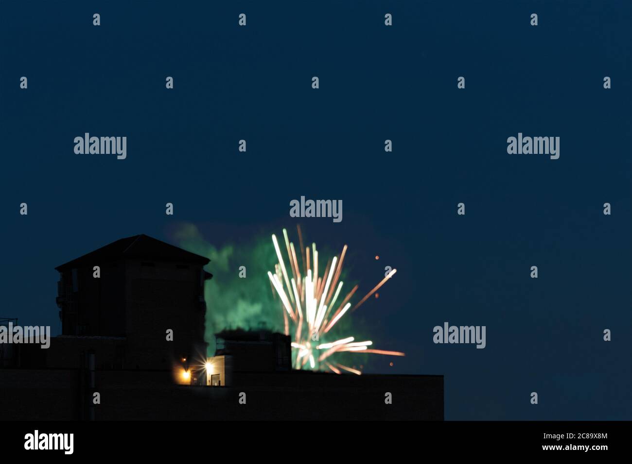 un'esplosione di fuochi d'artificio sul tetto di una città di new york, luce rosa e verde e fumo lampeggano dal tetto contro un cielo notturno blu scuro con copia Foto Stock